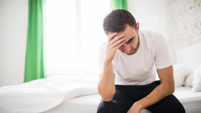 Ill man sitting on his bed with his head on his hand at home