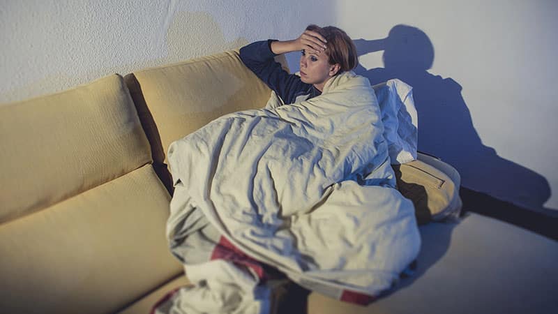 woman sitting on couch wrapped in duvet and blanket feeling miserable