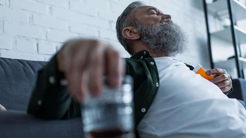 Bearded senior man with grey hair holding bottle with medication and glass of whiskey